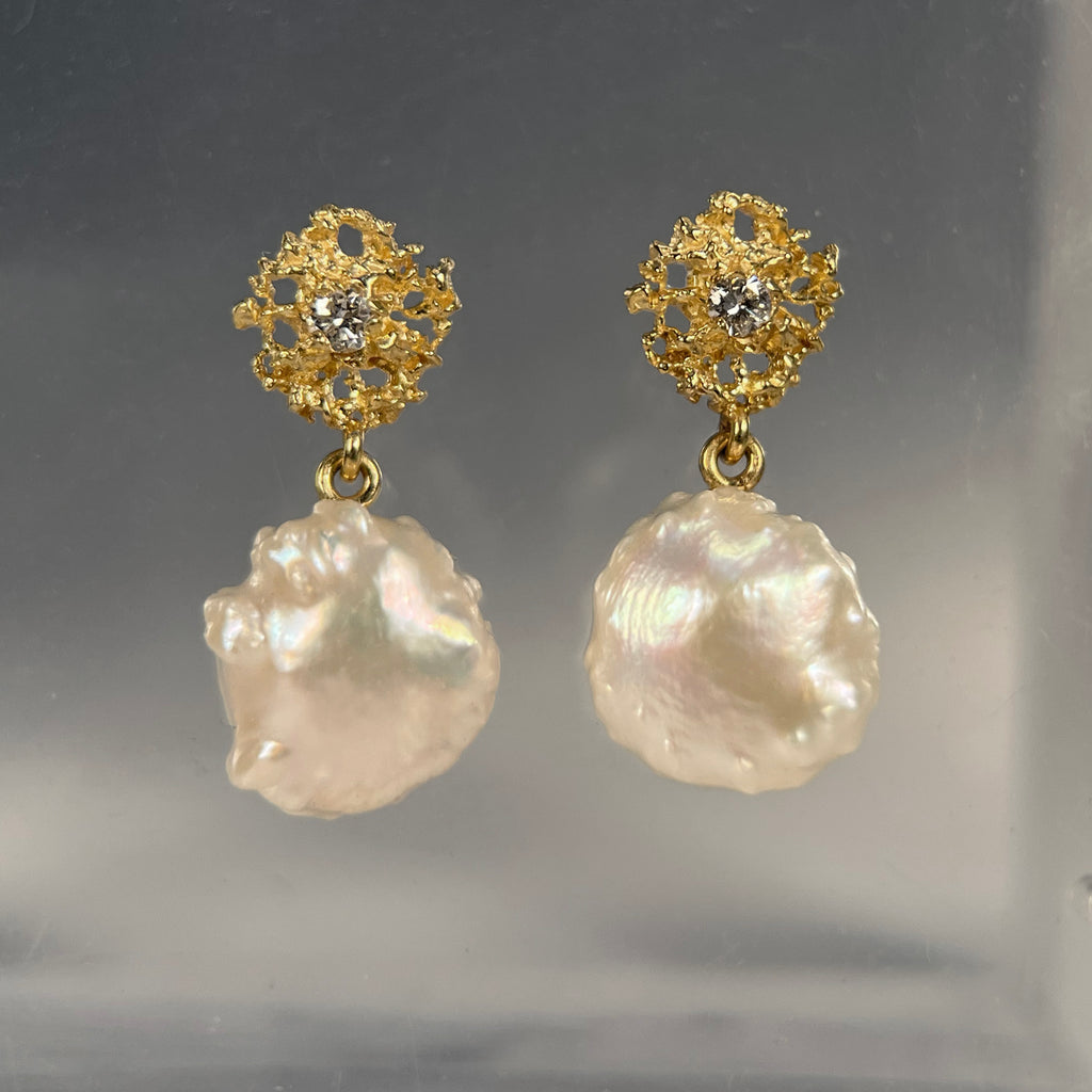 photo shows a pair of pearl earrings; the pearls are roundish but bumpy and irregular; they drop from a lacy, textured gold piece that is also roundish but smaller in diameter than the pearls and has a single diamond in the center 