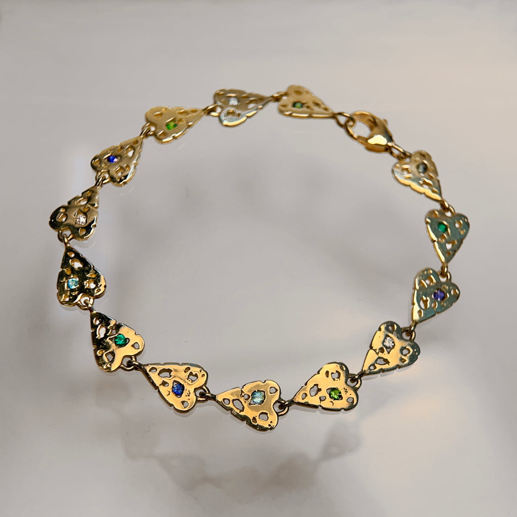 a bracelet made of 14 small hearts connected end to end; hearts are shown in 14k yellow, polished and with jagged holes; each heart is set with 1 stone, some blue and some green