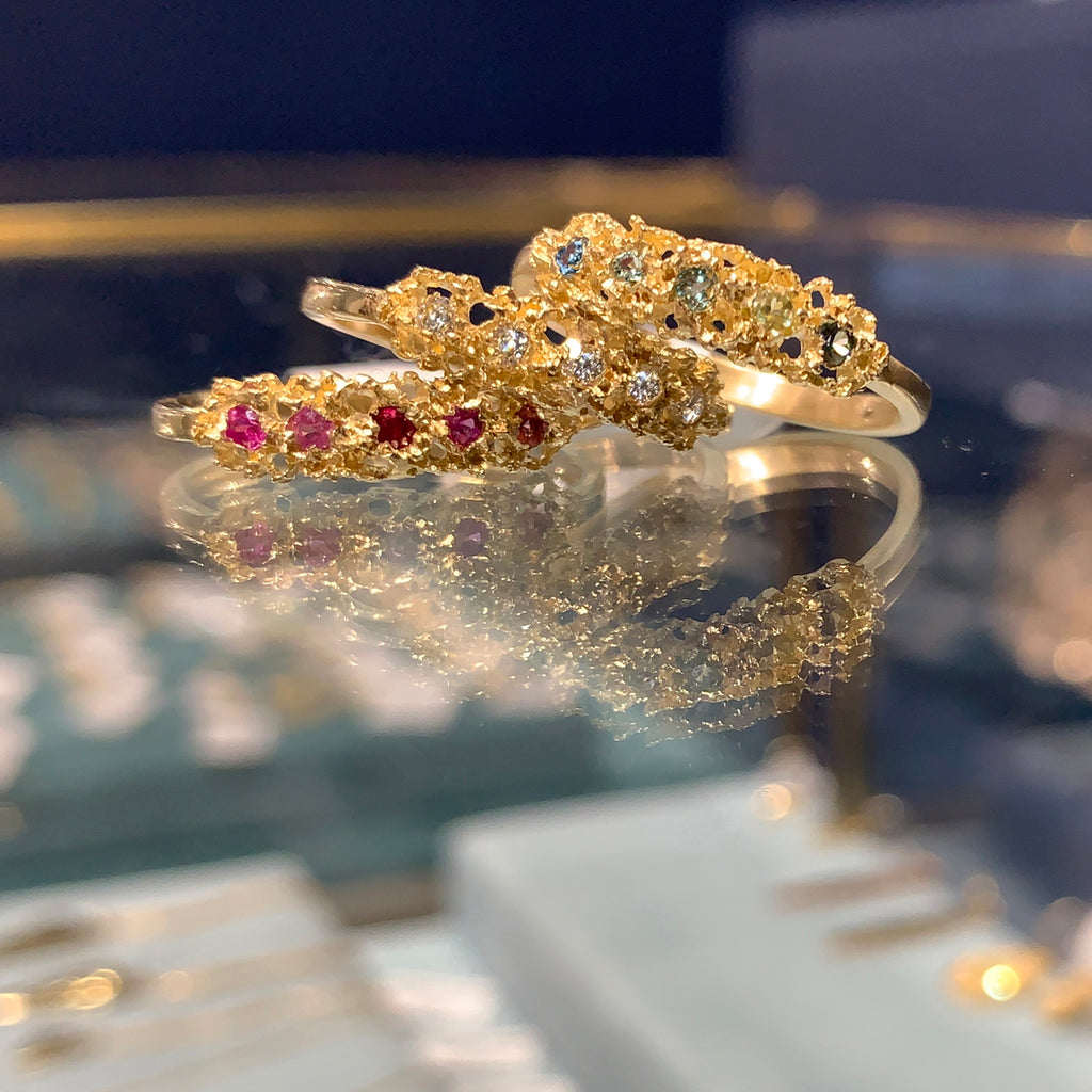 pile of 3 rings on glass case; shown in 14k yellow, with rubies, diamonds, and a mix of blue and green stones