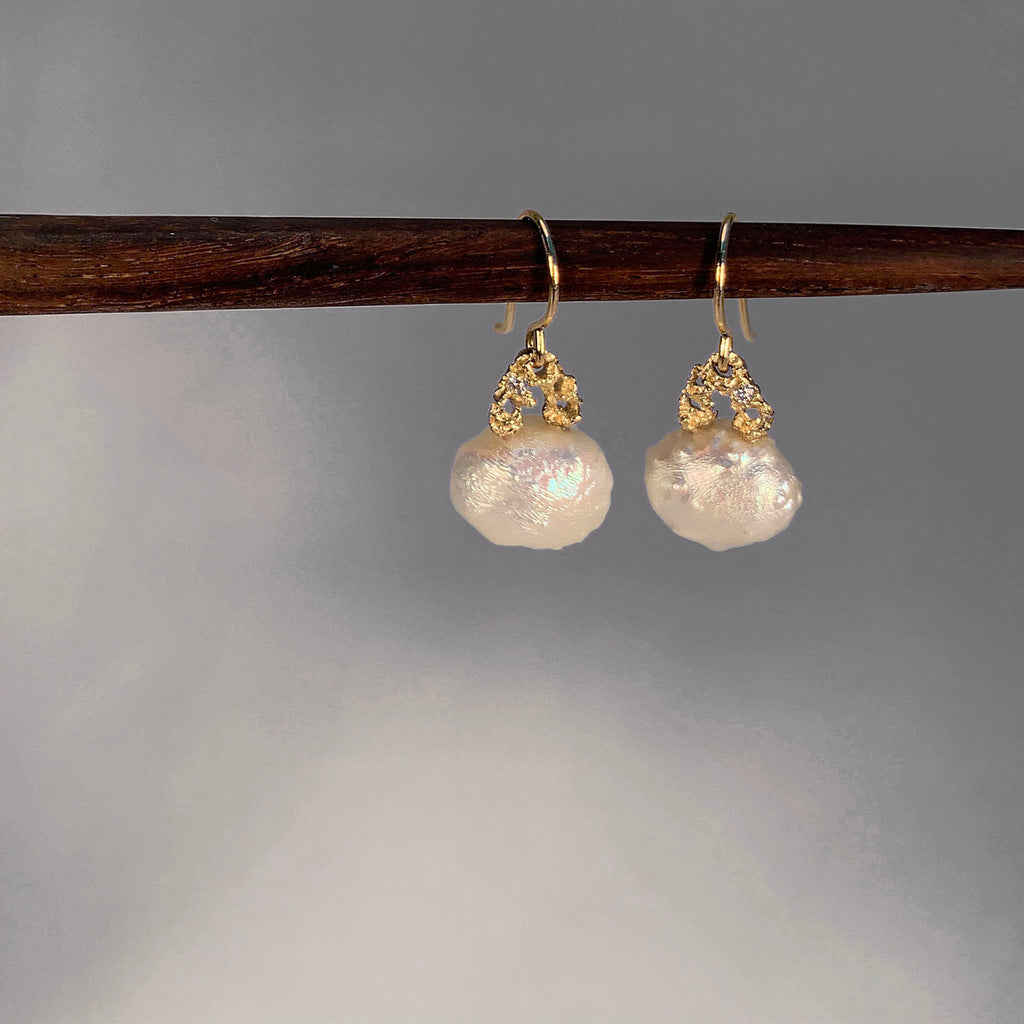A pair of small, bumpy pearl earrings on wires; each pearl is an imperfect horizontal oval, connected to a lacy gold arch with a single off-center diamond