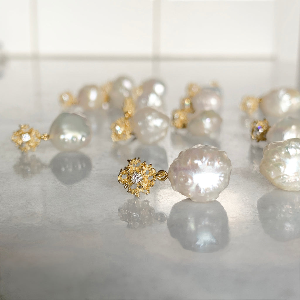 photo shows about 12 post earrings lying on their sides; the pearls are bumpy and irregular, but roundish; they drop from a lacy, textured gold piece that has a single diamond in the center 