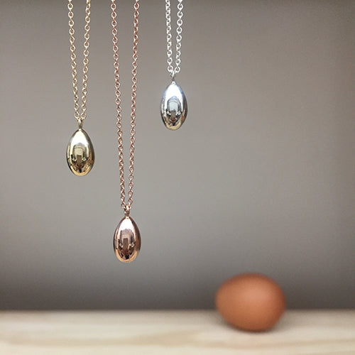 Three shiny egg necklaces, shown hanging and in silver, yellow gold, and rose gold. There is an out-of-focus real egg in the background.