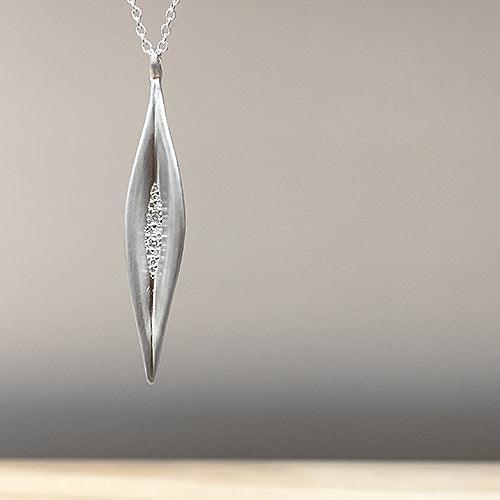 Slender, elongated, concave leaf shape base on a pod from the fruit of a magnolia tree; set with cluster of diamonds in various sizes in center