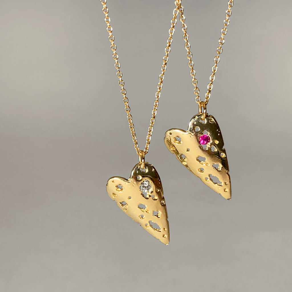 slightly curved, polished heart pendant with jagged holes throughout and a gemstone in the largest hole; 2 pieces shown in 14k yellow, one with a diamond and 1 with a ruby