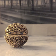 Small spherical box with tiny foot; tiny drops cover most of the top and bottom halves; a band of polished metal runs horizontally around the center where the box opens 